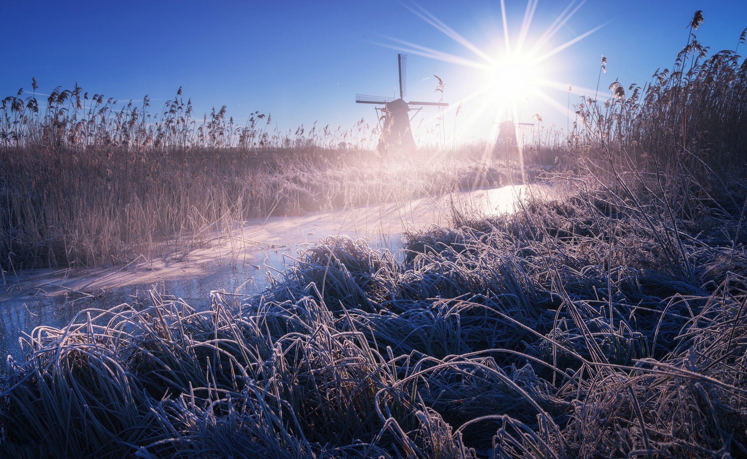 Download mobile wallpaper Winter, Nature, Sun, River, Windmill, Sunbeam, Man Made for free.